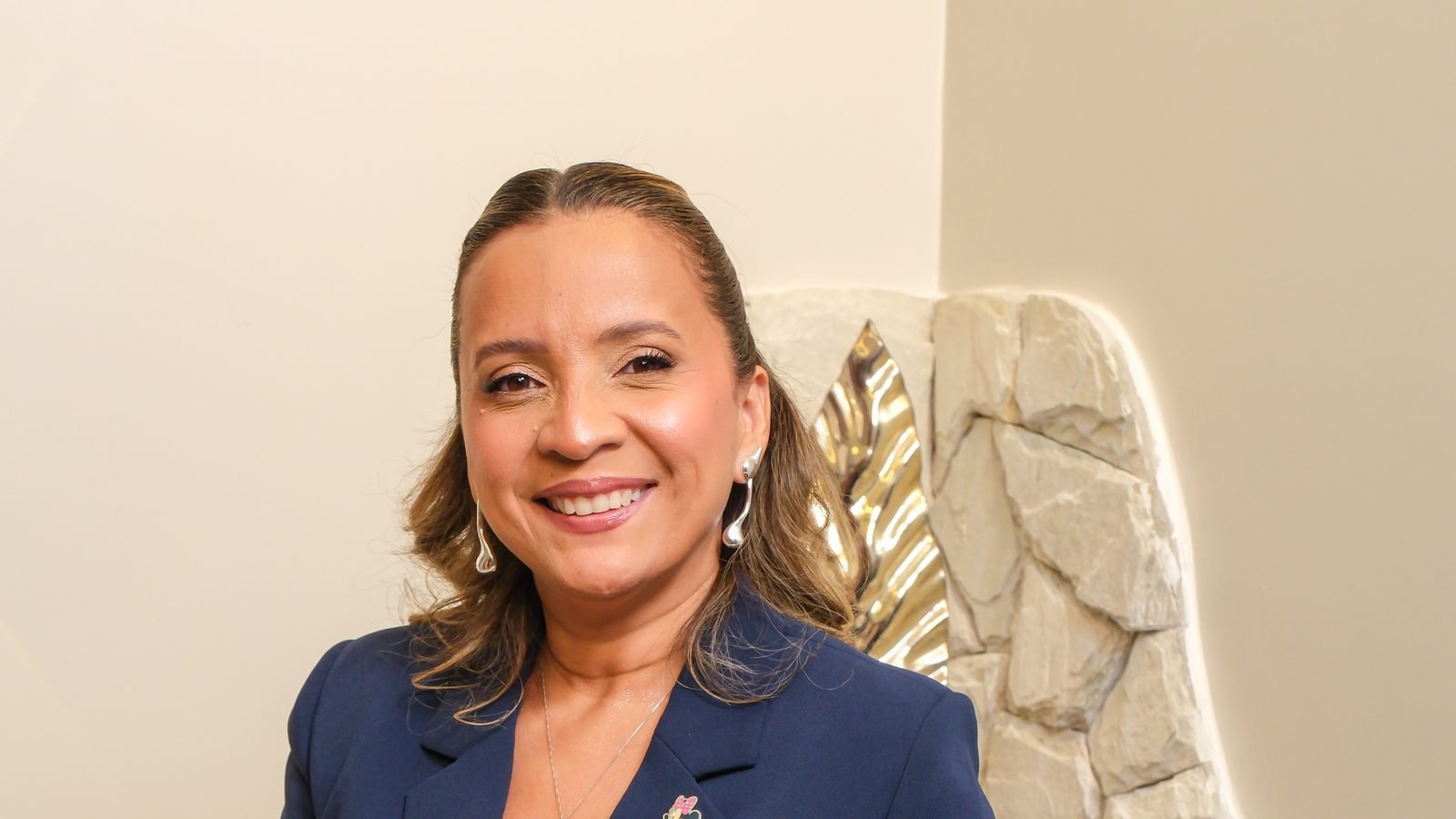 Kennya Martins, psicóloga e neuropsicóloga, sorrindo no consultório, com parede de pedra ao fundo