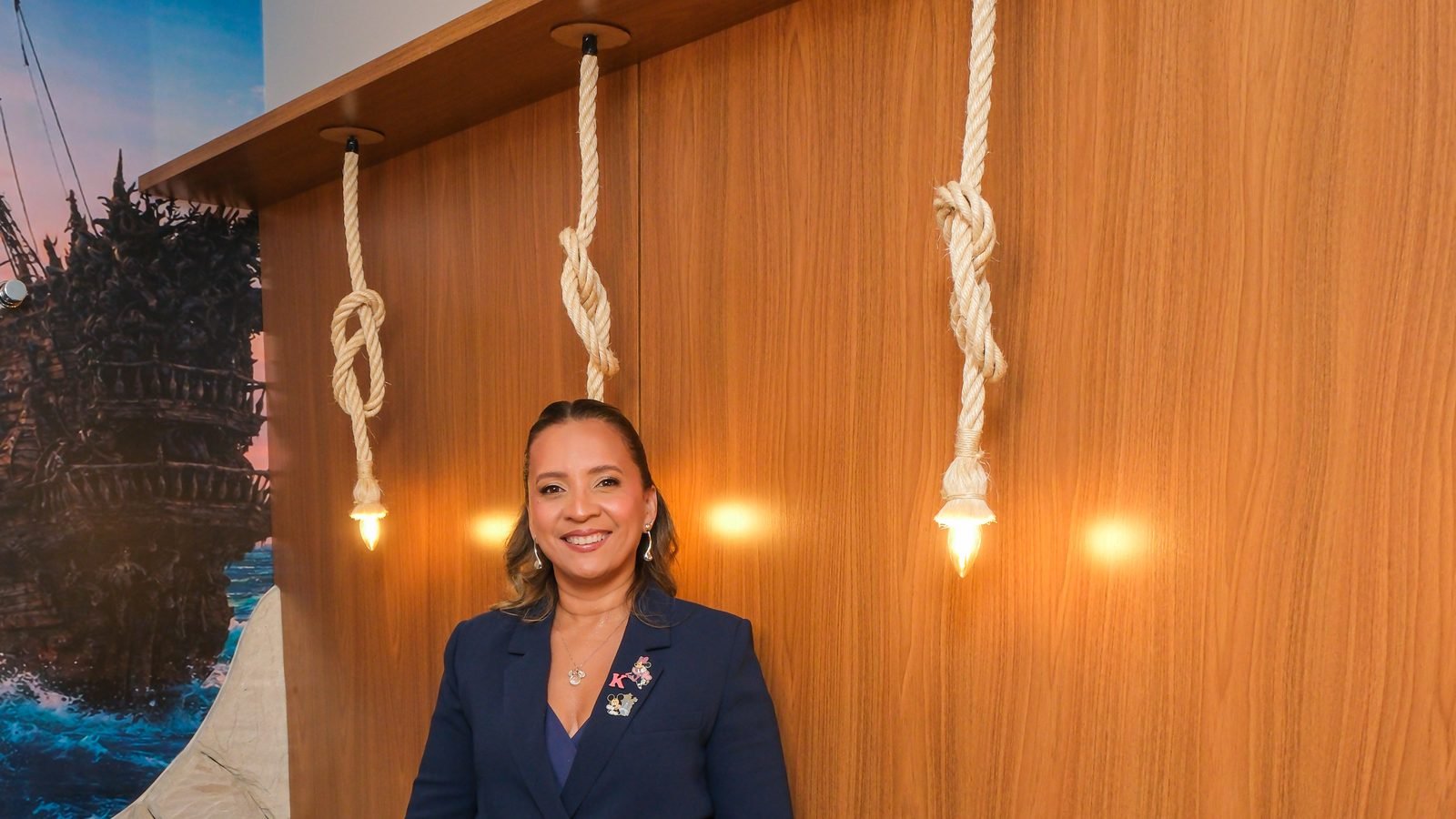 Kennya Martins, neuropsicóloga, diante de um painel de madeira com luminárias penduradas em cordas