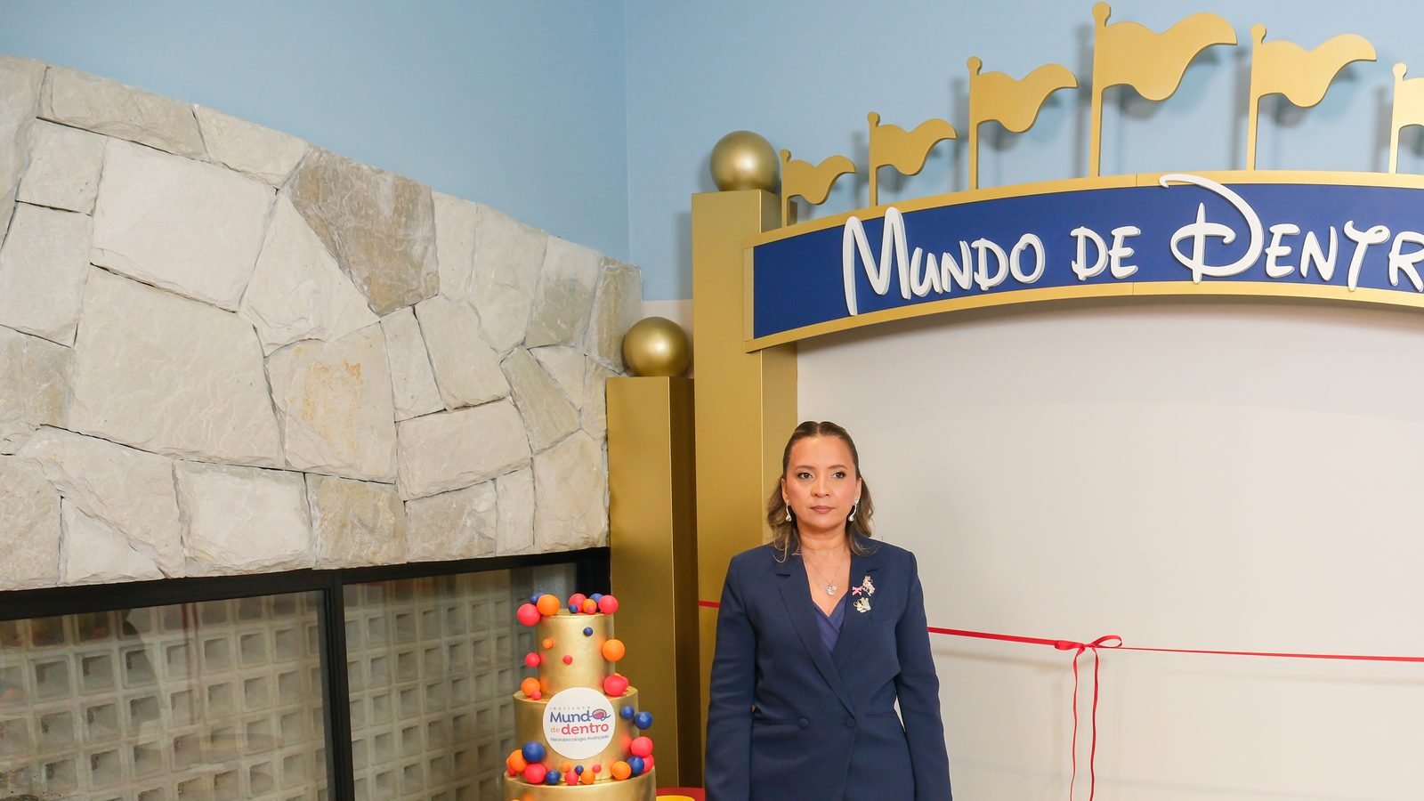 Kennya Martins ao lado do bolo de inauguração, com a parede de pedra do Instituto Mundo de Dentro ao fundo
