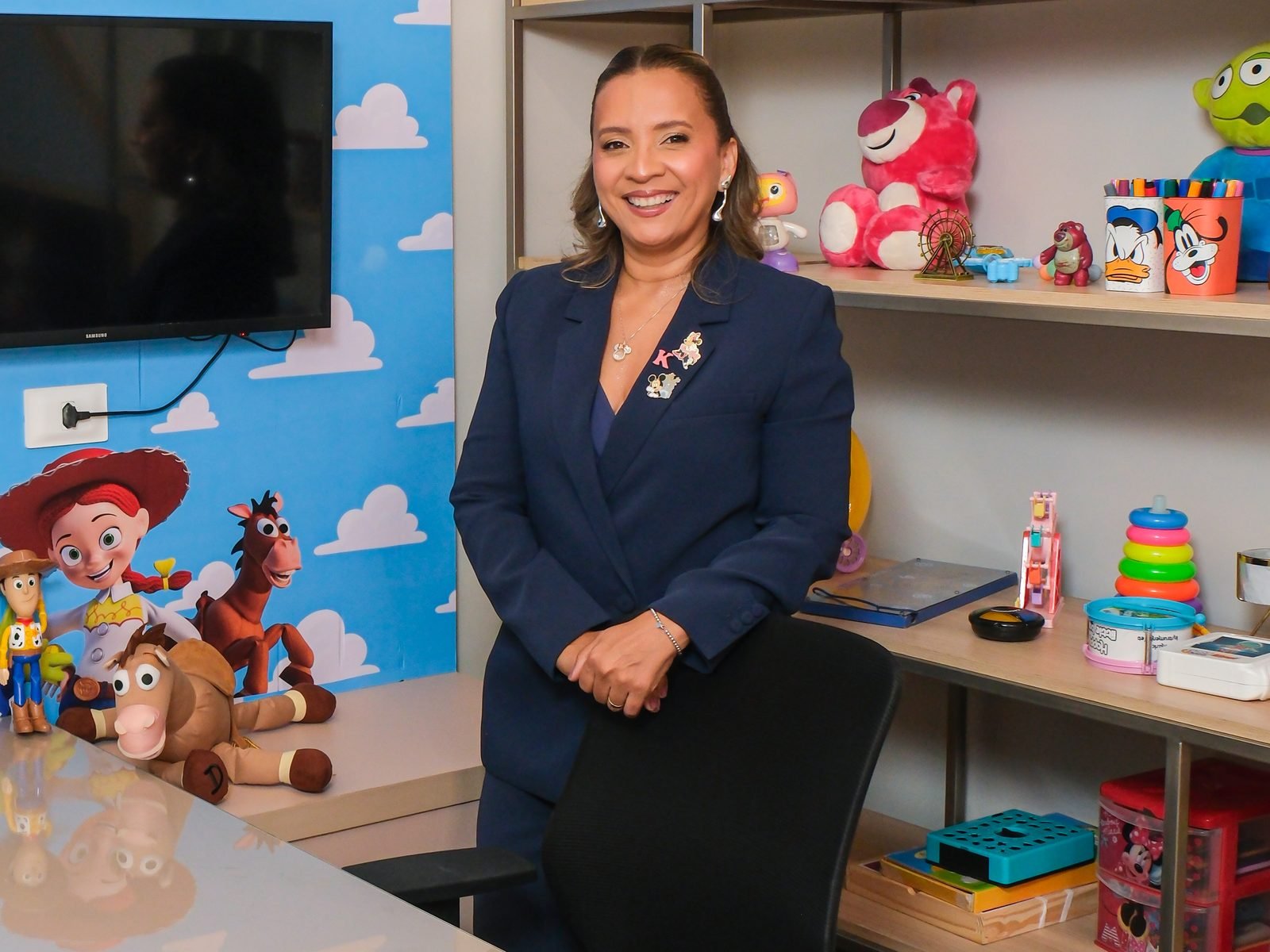 Kennya Martins sorrindo na sala lúdica, entre a mesa e a estante de brinquedos