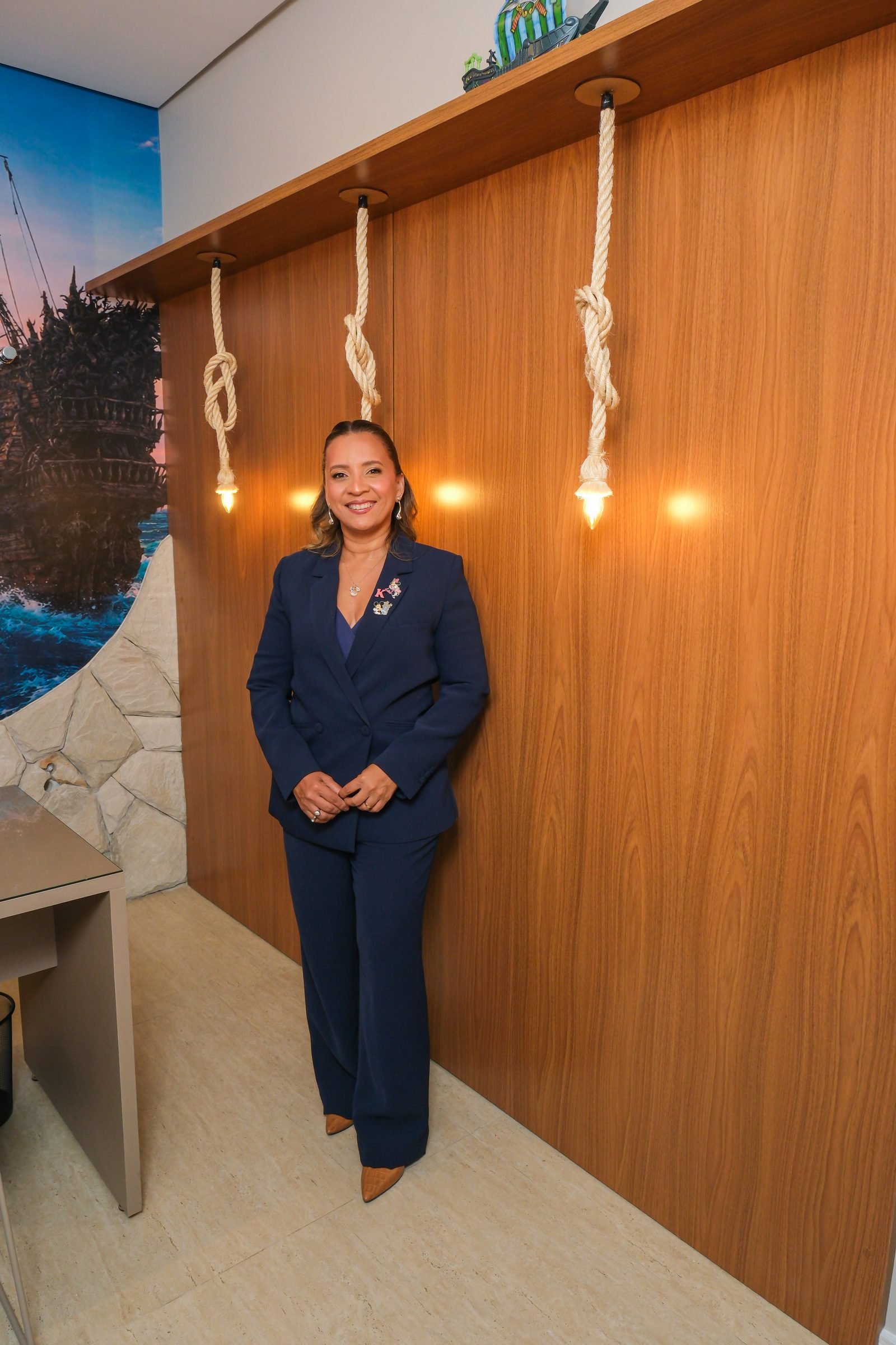 Kennya Martins, psicóloga, em retrato de corpo inteiro diante de um painel de madeira com luminárias de corda
